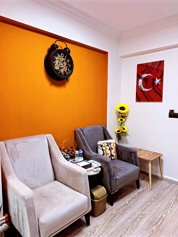 Turuncu duvarlı bekleme köşesi: iki koltuk, büyük bir saat ve Türk bayrağı.
